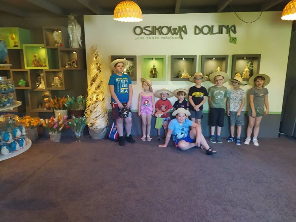 Grupa dziewczynek i chłopców w kapeluszach na tle wyrobów z osikowego drewna