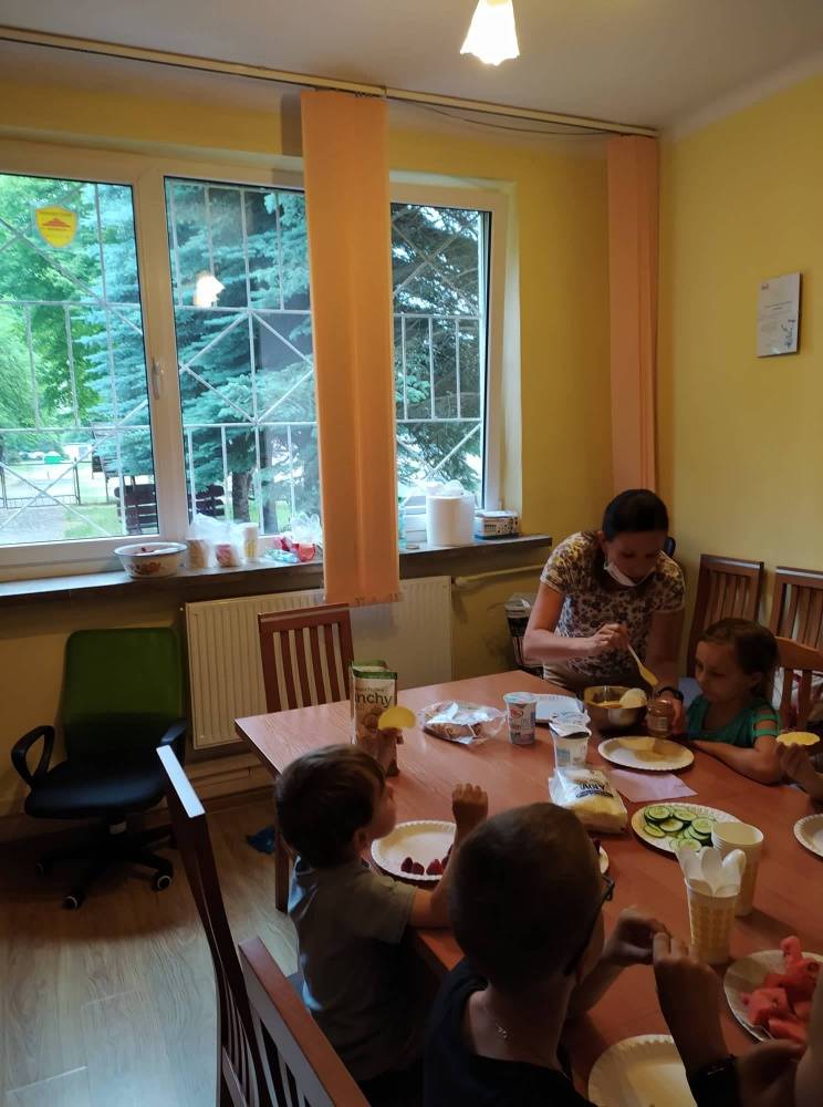 Grupa dzieci siedzi przy stole, na którym w pojemnikach poustawiane są pokrojone produkty spożywcze, a kobieta nakłada na talerzyk