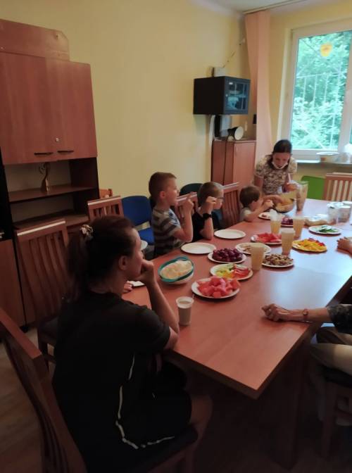 Grupa dzieci siedzi przy stole, na którym w pojemnikach poustawiane są pokrojone produkty spożywcze, a kobieta tłumaczy