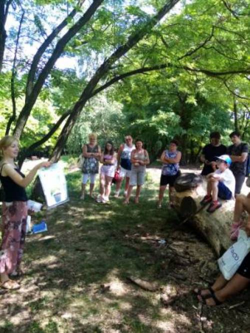 Liczna grupa osób stoi w parku przy drzewie i słucha wypowiadającej się kobiety