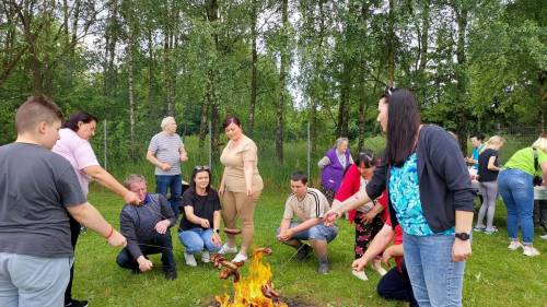 Grupa ludzi kuca i stoi przy ognisku i piecze kiełbaski na patykach. Są zadowoleni. Z tyłu widać stół i drzewa.