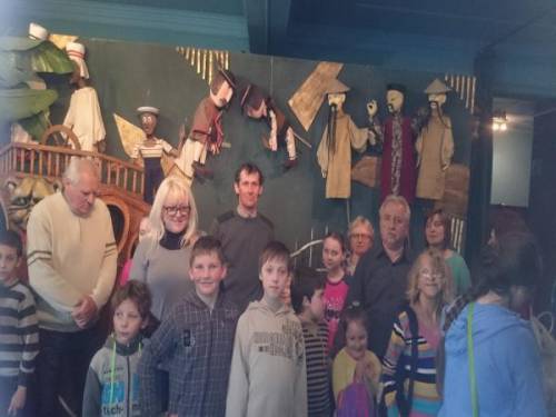 Grupa uczestników i uczestniczek projektu wraz z dziećmi stojąca we wnętrzu Teatru, a w tle kukiełki z przedstawień