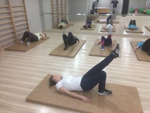 Grupa kobiet wykonuje ćwiczenia w sali gimnastycznej leżąc na plecach na macie