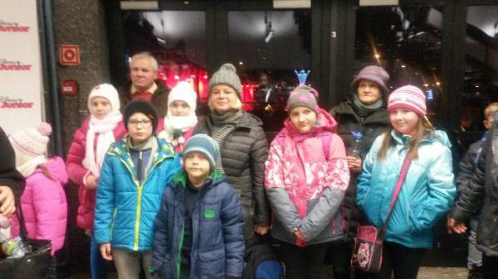 Grupa dzieci i dorosłych w kurtkach i czapkach stoi przed szklanymi drzwiami