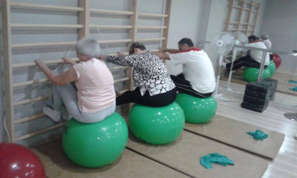 Trzy osoby siedzą na dużych piłkach twarzą do drabinek rehabilitacyjnych