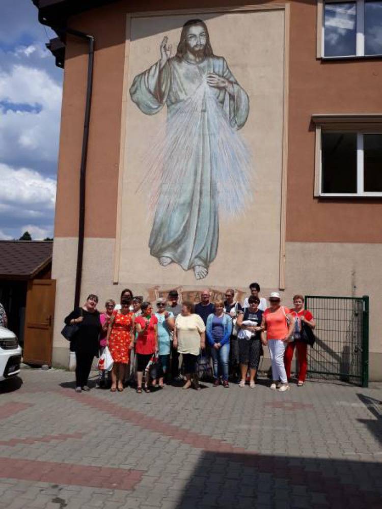 Grupa kobiet i mężczyzn stoi na tle budynku na którym znajduje się wizerunek Jezusa