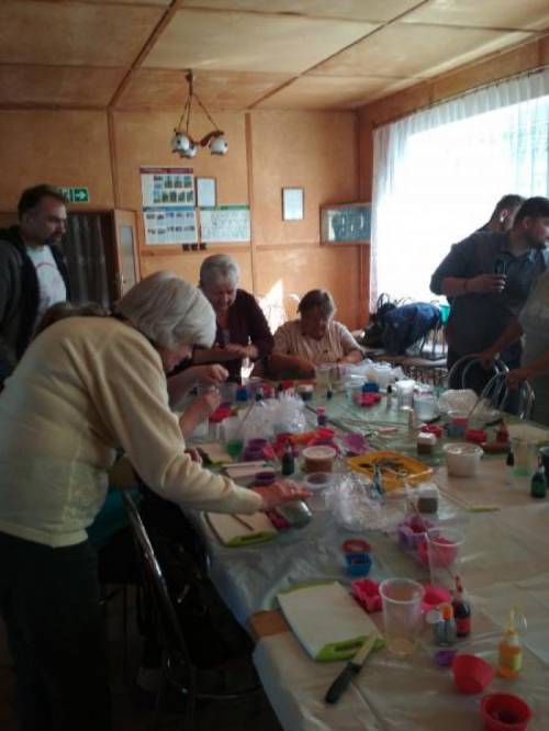 Kobiety i mężczyźni siedzą i stoją wokół stołu, na którym leży wiele plastikowych kubków, kolorowych foremek, fiolek, folii