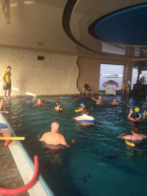 Grupa kobiet i mężczyzn w basenie wykonuje ćwiczenia z wykorzystaniem piankowych makaronów