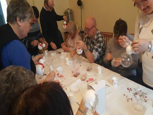 Grupa kobiet i mężczyzn przy stole maluje bombki w kształcie bałwanka