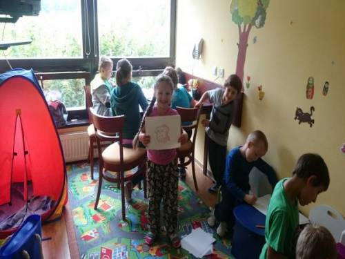 Dzieci uczestniczą w zajęciach plastycznych w sali, dziewczynka pokazuje swoją pracę
